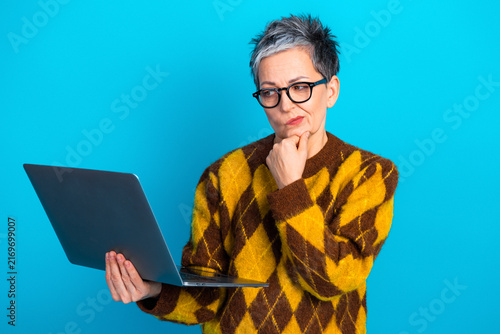 Mature woman with laptop looking thoughtful and puzzled while working remotely  wearing glasses and yellow argyle sweater against bright blue background