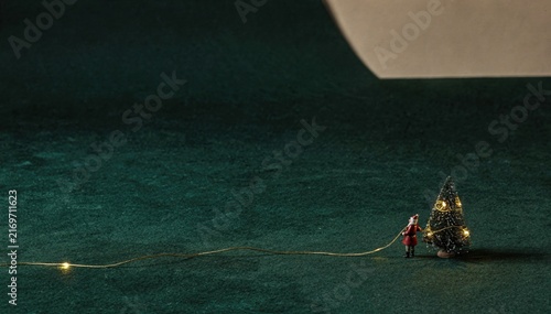 Vast deep-green field with a miniature person pulling a glowing gold light strand for a tiny Christmas tree, featuring generous copy space for holiday advertising