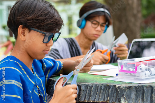 Two teenage boys in traditional shirts focusing on cutting paper with scissors for an art craft project together at an outdoor table.