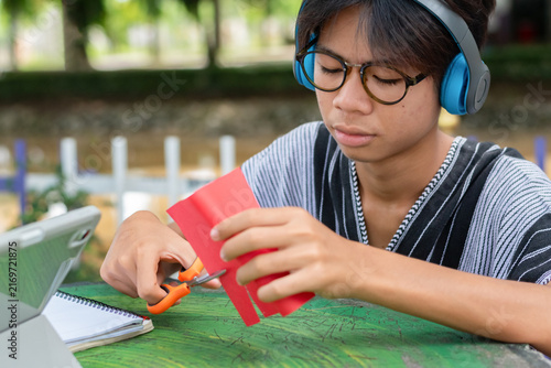 A young man with blue headphones and glasses cutting red paper with scissors outdoors, focused on an art craft project with a tablet nearby.