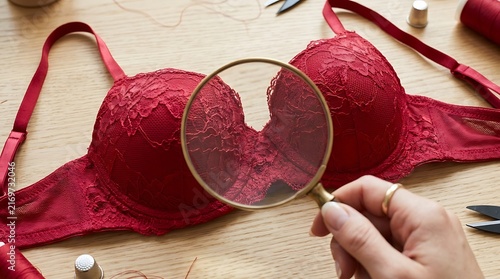 Hand holding magnifying glass over red lace bra on wooden table.