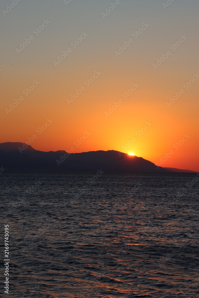 Fototapeta premium Beautiful orange sunset over the Corinthian Gulf in Loutraki, Greece, with the sun setting behind the distant Geraneia Mountains, creating a romantic and colorful Mediterranean seascape. 