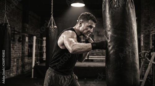 A muscular man in sleeveless shirt intensely trains with a punching bag in a dimly lit gym. Concept of men health, fitness, strength, sports training, and determination Generative AI