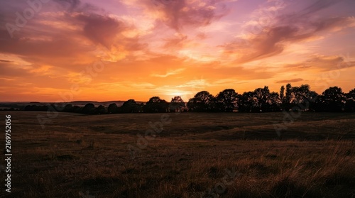 Vibrant sunset sky featuring warm orange purple gradient clouds over tranquil rural agricultural field with silhouette of tree line at horizon for nature landscape photography.