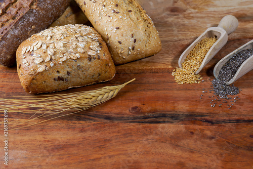 Assorted whole grain bread rolls, wheat stalk, and wooden scoops of chia and flax seeds on a rustic wooden background, promoting healthy eating
