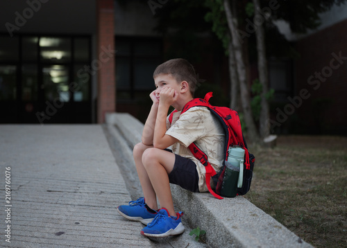 Childhood anxiety and school stress, lonely boy sitting outside with a backpack feeling sad and overwhelmed after class at the end of the school day.