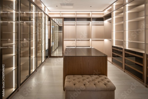 Modern luxury walk-in closet with integrated LED strip lighting, dark wood island, and tufted ottoman bench.