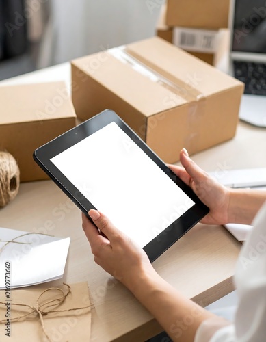 Hands holding digital tablet with blank mockup screen at desk with cardboard boxes for online store order management and shipping