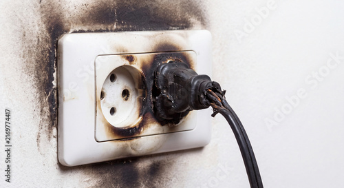 A dangerously burned and blackened electric wall socket with a melted power plug showing fire damage and soot on a residential plaster wall.