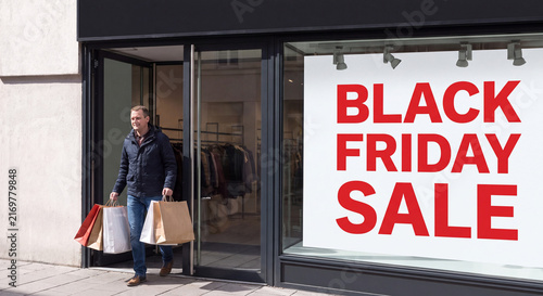 A stylish man holding multiple paper shopping bags walks past a glass retail storefront featuring a large Black Friday Sale sign