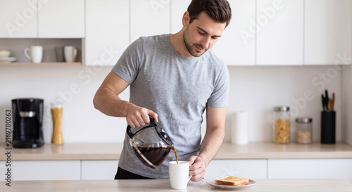 Young man standing in modern bright kitchen and pouring hot fresh coffee from glass pot into a white mug for breakfast