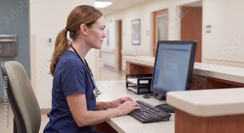 Female healthcare professional sitting at a hospital nursing station typing on desktop computer entering patient data