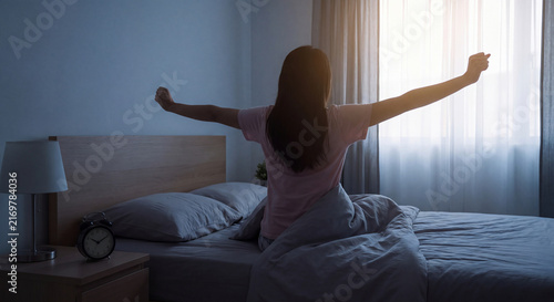 Back view of woman stretching both arms after waking up in comfortable bed during early morning at home