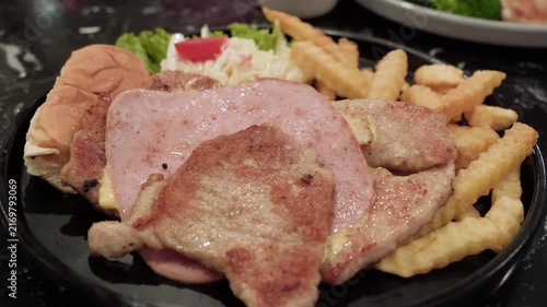 Close-up of a pork steak topped with ham and served with crispy French fries, bread, fresh lettuce, and creamy coleslaw on a black plate.