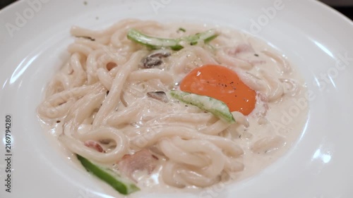 Creamy carbonara udon noodles served in a rich white sauce with mushrooms, bacon, and fresh cucumber slices, topped with a soft orange egg yolk and black pepper.