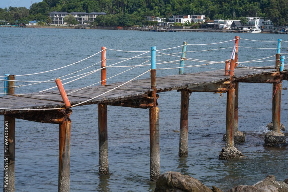 Fototapeta premium Weathered wooden walkway with rope railing extending above rocky coast in Thailand, connecting local waterfront area while overlooking tropical sea. Traditional tropical maritime landscape.