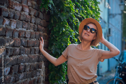 Fashionable young woman in sunglasses and straw hat posing by an old brick wall covered with lush ivy. Summer lifestyle portrait in warm evening light with urban charm and copy space