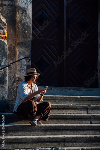 Solo female traveler relaxing on historic stone steps while using a smartphone at golden hour. Summer vacation, urban lifestyle, travel, sightseeing and digital connection