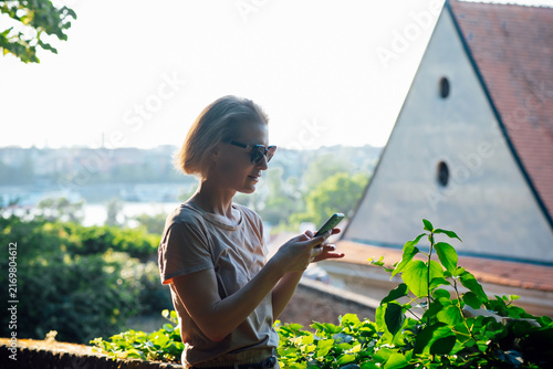 Young woman in sunglasses using a smartphone near historic rooftops at golden hour. Traveler exploring an old European town, checking maps and sharing vacation moments