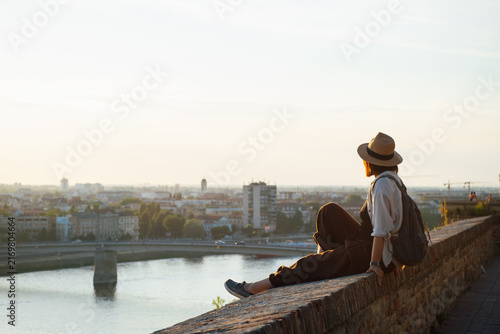 Young female traveler in hat and backpack relaxing on a historic fortress wall, admiring a panoramic river and city skyline at golden hour, capturing peaceful travel and summer adventure
