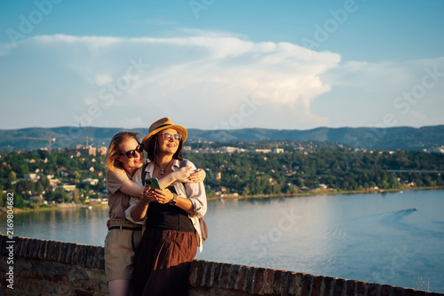 Two joyful female lesbian travelers taking a selfie at a scenic river viewpoint, hugging and waving during golden hour, celebrating friendship and love, travel and unforgettable summer moments