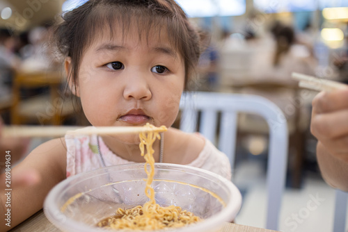 Konstfotografi Cute Asian Girl Eating Instant Noodles with Chopsticks at a Restaurant,Close-up of an adorable little Asian girl enjoying a bowl of delicious instant noodles with chopsticks in a casual restaurant