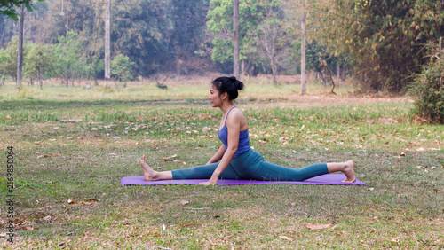 Young Asian woman practicing split yoga pose on mat in park. Healthy female stretching leg muscle, flexibility, outdoor fitness and wellness concept.