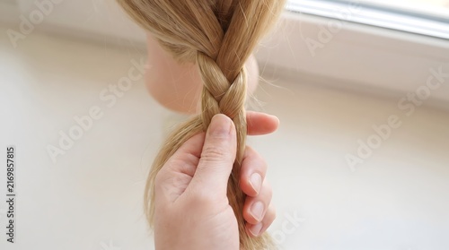 Hand braiding blonde hair on a training head, close-up of styling process