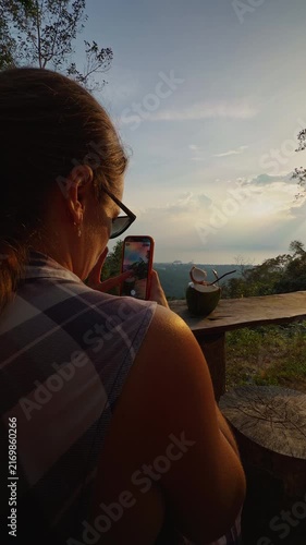 Woman Uses Phone to Photograph Fresh Coconut on Rustic Table During Sunset, Overlooking Forested Hills and Supporting Outdoor Travel Lifestyle Vertical video.