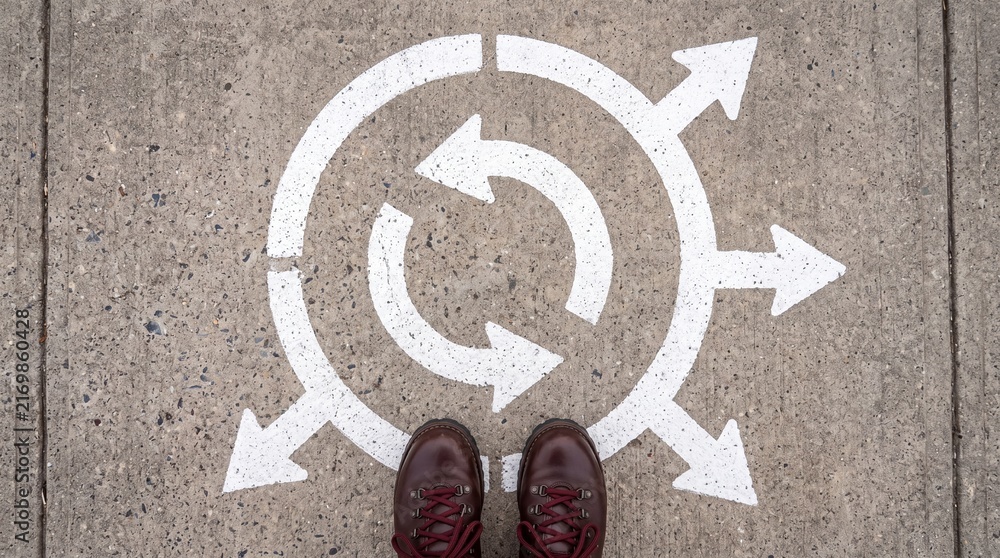 Naklejka premium A person standing at a crossroads with painted white arrows on the concrete pavement, symbolising choices, decisions and paths