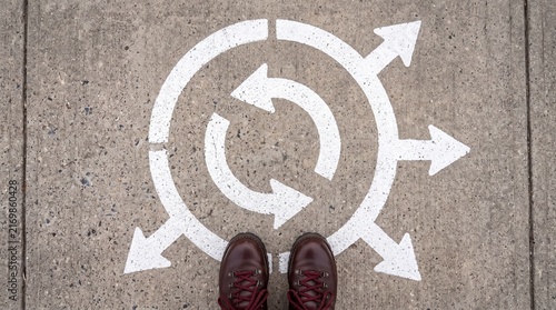 A person standing at a crossroads with painted white arrows on the concrete pavement, symbolising choices, decisions and paths