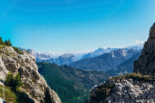 Forested slopes descend to a valley below snow tipped Dolomite peaks and clear blue sky. Panoramic alpine landscape and summer mountain travel for tourism and destination marketing