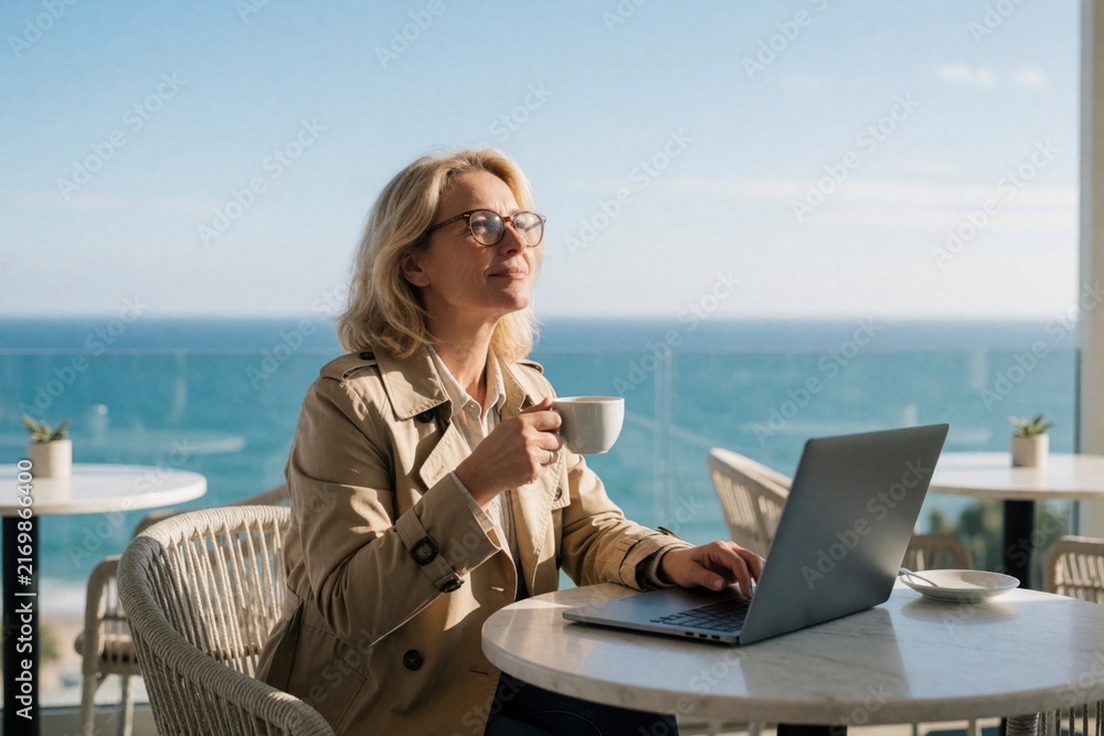 Obraz premium Woman works on laptop at cafe terrace with ocean view holding cup of tea. She looks inspired and relaxed, enjoying sunny spring day, dreaming of success and future projects.