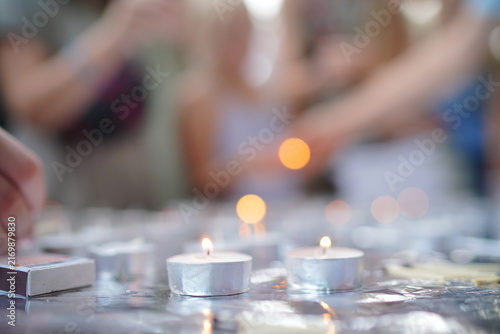 Фотография Softly lit tealight candles glow on a reflective surface as blurred people reach to light them, creating a peaceful, reflective atmosphere for memorials, vigils, prayer, remembrance and quiet tribute