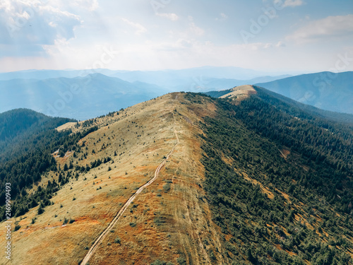 Aerial panorama of a long grassy mountain ridge with a winding hiking trail, surrounded by forested slopes and distant blue mountains.