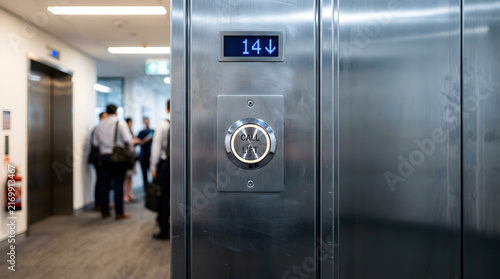 Modern stainless steel elevator call lift button with illuminated ring and blue digital floor indicator in a busy corporate office building lobby with professional people blurred in background