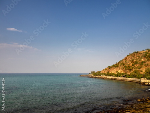 View of the turquoise sea meeting the rocky coastline with sparse vegetation on the hill under a clear blue sky in Ras Al-Khaimah, Ras Al Khaimah, United Arab Emirates.