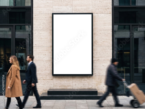 Vertical outdoor poster mockup mounted on a stone building facade with blurred pedestrians walking along a city street for brand design showcase.