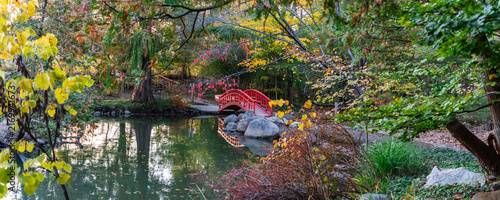 Aerial view of a red arched bridge spanning a tranquil pond in a lush park with autumn foliage and surrounding trees in Troy, Michigan, United States.