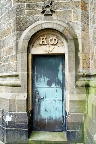Weathered bronze door set within a stone church wall. The arch above displays the alpha and omega symbols carved in relief. 