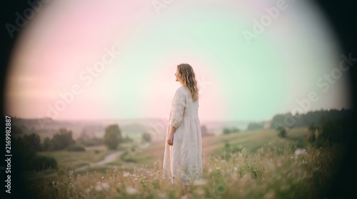 Woman standing in a grassy field at sunset wearing a long linen dress, captured with a soft vignette and dreamy pastel light atmosphere