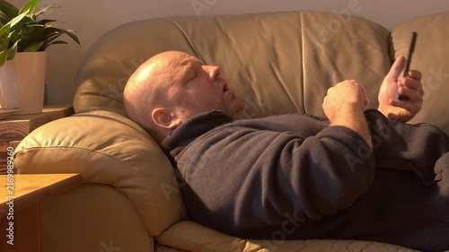 Middle-aged white man laying comfortably on a tan coloured leather sofa, doom scrolling on the cellphone, sneezes, and continued to scroll through the phone.