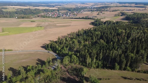 Smooth aerial view over lush green countryside with open fields, dense trees, winding rural roads and peaceful natural scenery in bright daylight.