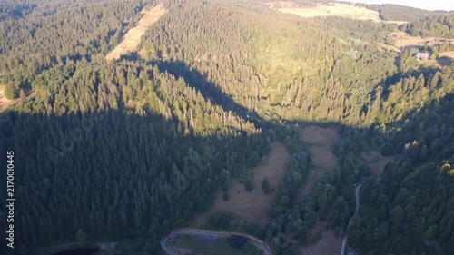 Smooth aerial view over lush green countryside with open fields, dense trees, winding rural roads and peaceful natural scenery in bright daylight.