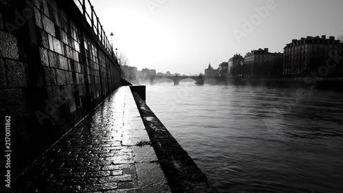 Black and white view of a misty river with a stone wall and bridges in the distance during a tranquil day.