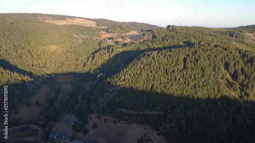 Smooth aerial view over lush green countryside with open fields, dense trees, winding rural roads and peaceful natural scenery in bright daylight.