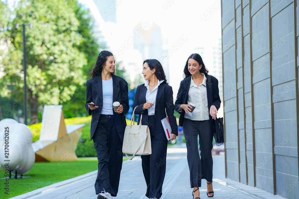 Obraz premium A team of three Hispanic businesswomen in matching dark suits, laughing openly as they walk, walk three abreast along a sunlit avenue carrying tote bags, folders and phones.