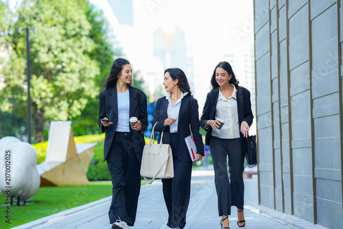 A team of three Hispanic businesswomen in matching dark suits, laughing openly as they walk, walk three abreast along a sunlit avenue carrying tote bags, folders and phones.