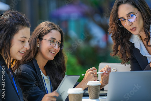 Quadro su tela A team of three Hispanic businesswomen in matching dark suits, deep in an animated discussion, sketch out an idea with a stylus on a tablet, part of a women in business series