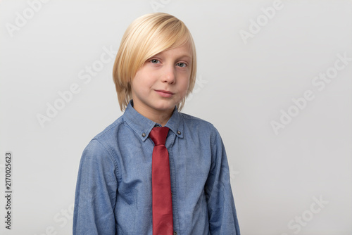 Young boy with blonde hair and a red tie looking directly at the camera, conveying a sense of smartness and formal confidence on a light background